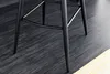 Close-up of the legs of a black bar stool on a dark wooden floor, with a yellow rug partially visible. Produktbild-Vorschau