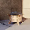 A fire pit with a mesh lid and a tool on a concrete surface, with a stone wall in the background. Produktbild-Vorschau