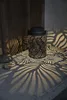 A black metal lantern with a leaf pattern is casting shadows on a concrete surface and a wooden wall. Produktbild-Vorschau