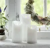 Three white LED candles of varying heights are arranged on a surface in front of a window with a floral wreath. Produktbild-Vorschau