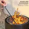 A hand is using a poker to tend a fire in a black metal fire pit. The fire pit is outdoors, with gravel and a textured wall in the background. Text overlays provide product information. Produktbild-Vorschau