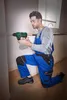A man in blue overalls and safety glasses is kneeling and using a green and black cordless drill to drill into a wall. Sawdust is visible on the floor. Produktbild-Vorschau