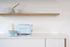 A light blue Smeg toaster sits on a white countertop next to a blue bowl and a cream-colored bottle. A wooden shelf with two bowls is above. Náhled obrázku produktu