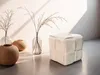 A beige pouf sits on a gray floor in front of a white wall, with decorative vases and dried plants. Produktbild-Vorschau
