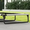 A close-up view of a raised dog bed with a gray fabric surface and black metal legs, set on a patio with a grassy area and bushes in the background. Produktbild-Vorschau