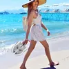 Hellgraue Stoffsandalen mit Schleifendetails, gehalten von einer Frau, die an einem Sandstrand entlanggeht Anteprima dell’immagine prodotto