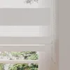 A close-up view of a white roller blind with a cassette, showing its textured fabric and control chain, with a blurred view of trees and sky visible through the window below. Produktbild-Vorschau