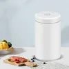 A white vacuum sealer sits on a table next to a cutting board with raw meat and vegetables, and a bowl of produce. Produktbild-Vorschau