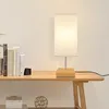 A table lamp with a fabric shade sits on a wooden shelf next to a stack of books. Náhľad obrázku produktu