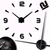 A close-up view of a large, modern, DIY wall clock with black numbers and hands against a white background. The clock mechanism is partially visible in the bottom left corner. Produktbild-Vorschau
