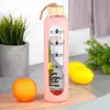 A pink glass water bottle with time markings and a silicone sleeve, placed on a wooden cutting board with an orange and lemons in the background. Voir les images du produit