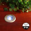 A round, silver LED ground light is embedded in a wooden deck, surrounded by green ivy leaves. The light is illuminated and shows water droplets on its surface. A circular icon with a cloud and rain, and the text 'IP 44' is in the bottom right corner. Produktbild-Vorschau