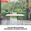 A white folding table is set up on a wooden deck outdoors, with a garden and trees in the background. Produktbild-Vorschau