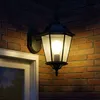 A black outdoor lantern with a bright white LED bulb is mounted on a brick wall next to a white-framed window. Náhľad obrázku produktu