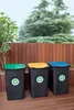 Three recycling bins with different colored lids (green, yellow, blue) are placed on a wooden deck in front of a wooden fence and trees. Produktbild-Vorschau