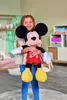 A young girl is holding a large Mickey Mouse plush toy in a bedroom setting. Voir les images du produit