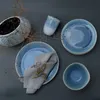 A collection of blue ceramic tableware, including plates, a bowl, and a cup, arranged on a dark gray surface with dried plant sprigs and scattered salt crystals. Náhľad obrázku produktu
