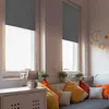 A cozy room with two windows covered by gray blackout roller blinds, a sofa with colorful pillows, and a white cabinet with a lamp and a mug. Anteprima dell’immagine prodotto