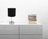 A black fabric lampshade table lamp with a silver base sits on a white cabinet next to a stack of books. Produktbild-Vorschau