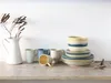 A collection of ceramic tableware, including mugs and plates, arranged on a wooden surface against a white wall. Náhľad obrázku produktu