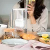 A person is drinking from a glass of water next to a water filter pitcher, croissants, and a jar of nuts on a table. Produktbild-Vorschau