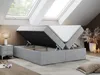 A bedroom with skylights, featuring a grey upholstered bed frame with its storage compartment open, revealing the mattress inside. A nightstand with a lamp is visible to the left, and decorative plants and candles are on the right. Produktbild-Vorschau