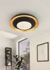 A modern ceiling light fixture with a black and gold circular design is mounted on a white ceiling in a room with a window and framed artwork. Náhled obrázku produktu