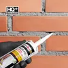A hand wearing a black glove is holding a tube of sealant, applying it to a brick wall. Produktbild-Vorschau