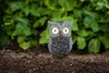 A small, gray owl figurine is placed on dark soil in front of a blurred green leafy background. Produktbild-Vorschau