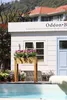 A raised garden bed filled with flowers sits in front of a white building with a blue door and window, next to a pool. Anteprima dell’immagine prodotto