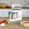 A white stand mixer with a stainless steel bowl is displayed on a wooden countertop, with a plate of berries and cream and a plate of cake in the foreground. The background shows a kitchen counter with cabinets and a backsplash. Anteprima dell’immagine prodotto