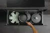 A hand is reaching into an open kitchen drawer to take out a frying pan. The drawer contains pots and a protective mat. Produktbild-Vorschau