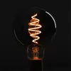 A close-up shot of a vintage-style LED light bulb with a spiral filament, glowing with warm light against a dark background. Produktbild-Vorschau