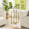 A round side table with a marble-look top and gold metal legs is placed between two cream-colored armchairs, next to a large potted plant. A window with green foliage is visible in the background. Produktbild-Vorschau