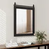 A rectangular mirror with a black frame and a sliding barn door mechanism is mounted on a white paneled wall above a wooden console table. On the table, there are books, a candle, a pair of glasses, and a vase of pink tulips. Podgląd zdjęcia produktu