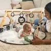 A baby is playing on a playmat under a wooden gymini with toys, with an adult nearby. Produktbild-Vorschau