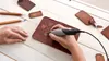 A person is using a Dremel Stylo+ rotary tool to engrave a compass design onto a brown leather item, possibly a wallet or luggage tag. Several other leather items and carving tools are visible on a white wooden surface. Anteprima dell’immagine prodotto