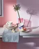 A still life arrangement of teacups, saucers, and a flower on a table, with a split pink and purple background. Náhled obrázku produktu
