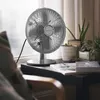 A silver desk fan sits on a shelf next to potted plants and magazines, with sheer curtains in the background. Produktbild-Vorschau