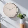 A beige wall clock with embossed numbers is mounted on a white wall, with shadows cast on the wall. A small white vase with green foliage and white flowers sits on a wooden stand to the right of the clock. Anteprima dell’immagine prodotto