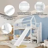 A white bunk bed with a house-shaped top, a slide, and stairs with storage compartments, set in a child's bedroom. Produktbild-Vorschau
