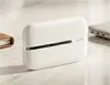A white mobile router is placed on a marble-like surface next to a notebook and a laptop. Produktbild-Vorschau