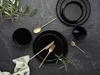 A black stoneware dinnerware set with gold cutlery is arranged on a dark, textured surface, accented by green foliage. Produktbild-Vorschau