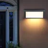 An outdoor wall light illuminates the wall next to a dark front door with decorative glass panels. Náhled obrázku produktu