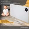 A baby sitting on a wooden floor next to a white infrared heater, with marketing text overlays. Produktbild-Vorschau