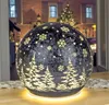 A decorative LED sphere with a Christmas tree and snowflake design, illuminated from within, sits on a table in front of a blurred background of a house and a Christmas tree. Produktbild-Vorschau