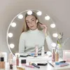 A young woman is applying makeup in front of a lighted vanity mirror, with various makeup products arranged on the vanity table. Náhled obrázku produktu