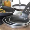 A stack of frying pans on a wooden surface, with a yellow bell pepper and some vegetables in the background. Produktbild-Vorschau