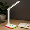 A white LED desk lamp with a colorful base light is shown on a wooden desk next to an open notebook and a pencil. A potted plant is visible in the background. Produktbild-Vorschau