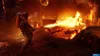 A person in a hazmat suit uses a flamethrower in a burning building, with bodies on the ground. Produktbild-Vorschau